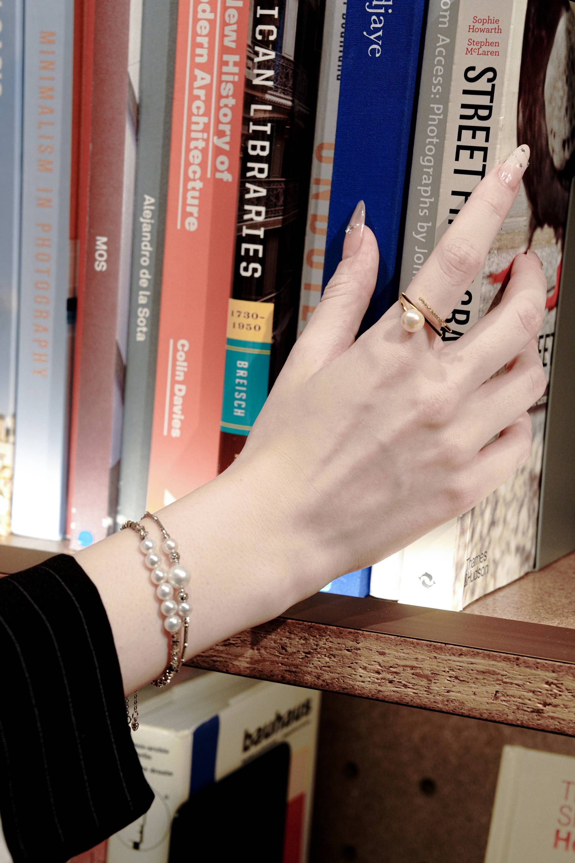 Intellectual chic lifestyle shot of a woman browsing a bookshelf, wearing Cyrene's layered pearl bracelets for a refined everyday look