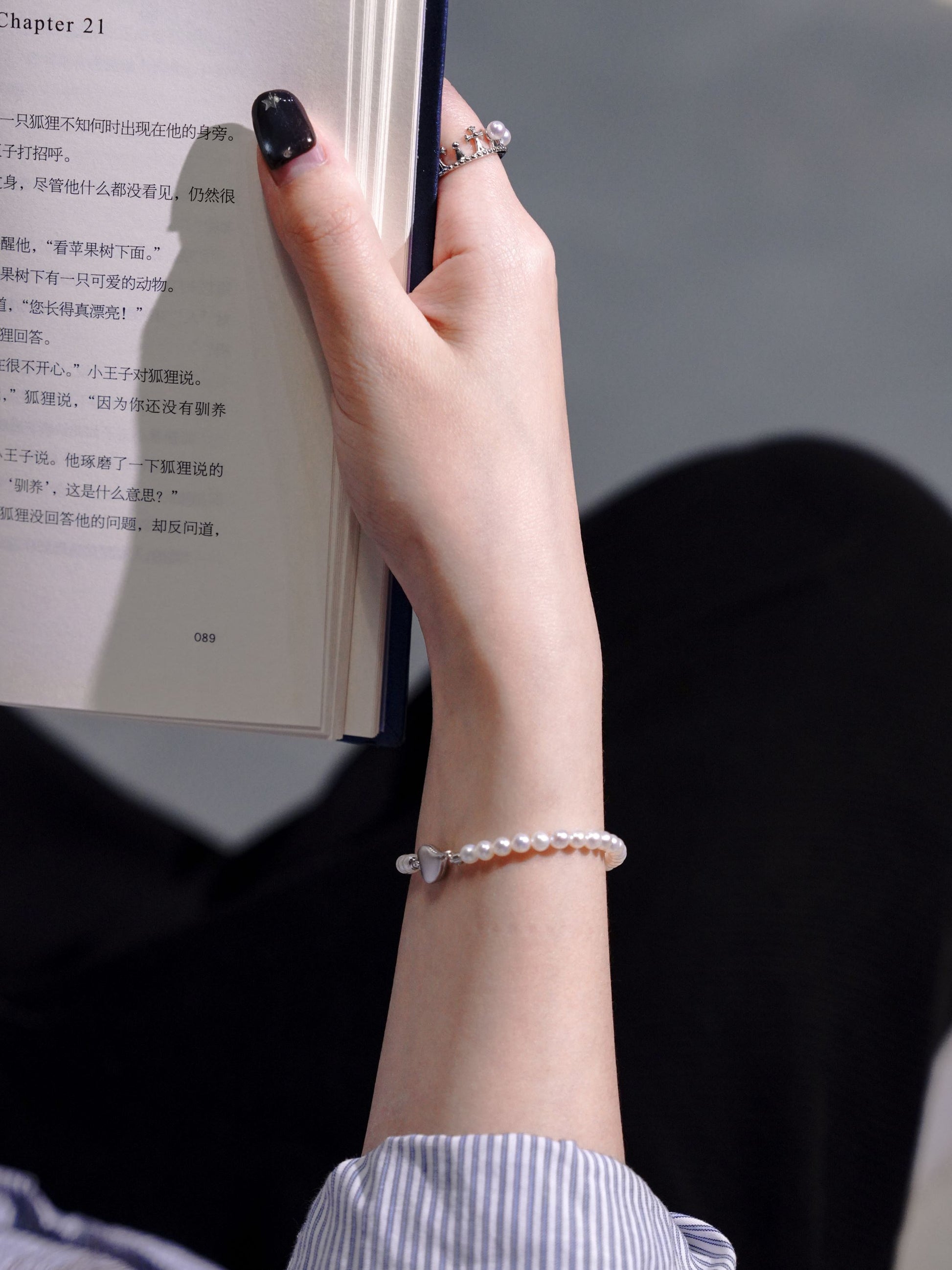 Lifestyle shot of a woman holding a book, elegantly accessorized with Cyrene's delicate seed pearl bracelet with a heart clasp.