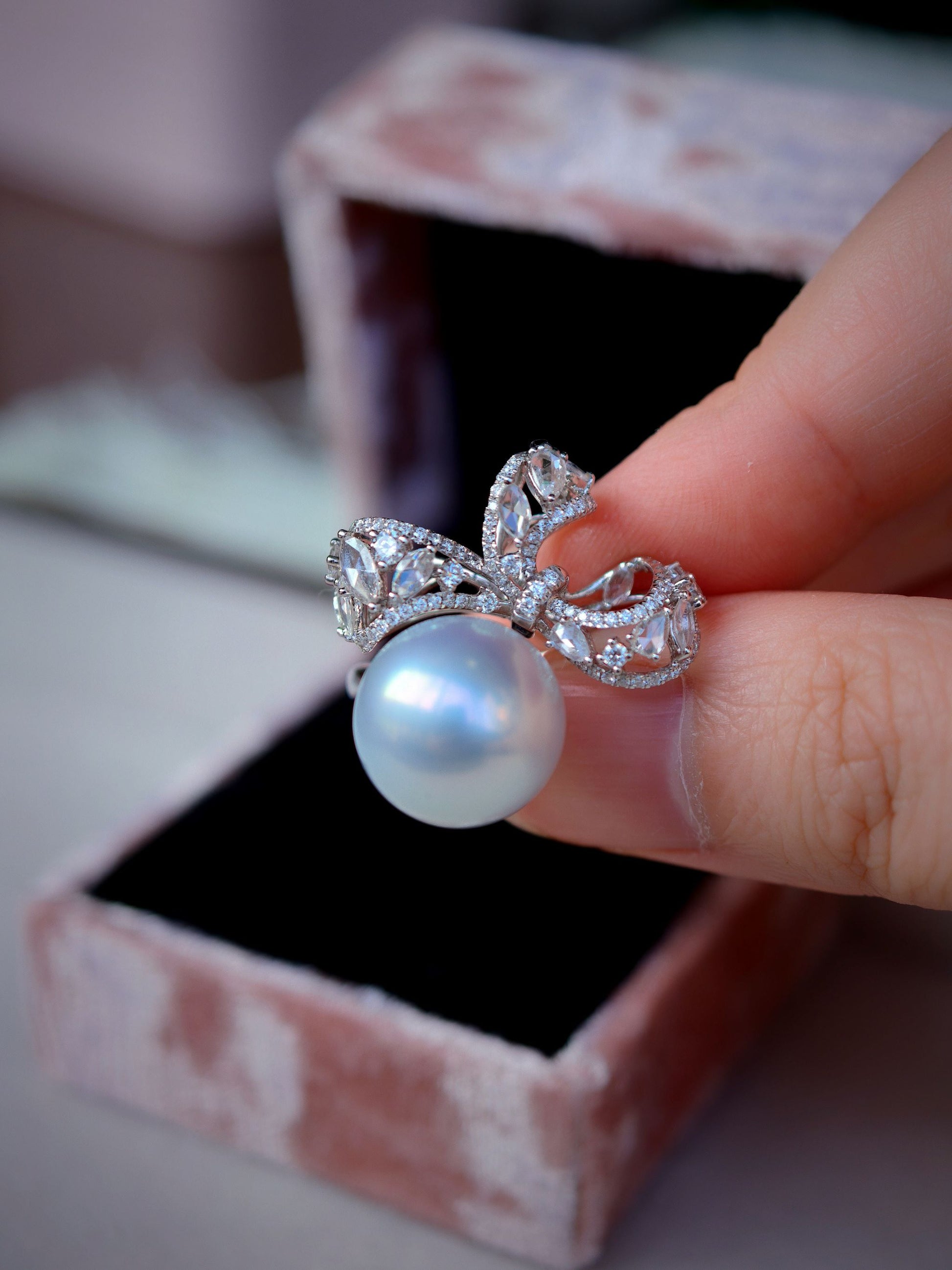 Cyrene Solitaire Bow Pearl Ring held between fingers above gift box — South Sea pearl and diamond ring